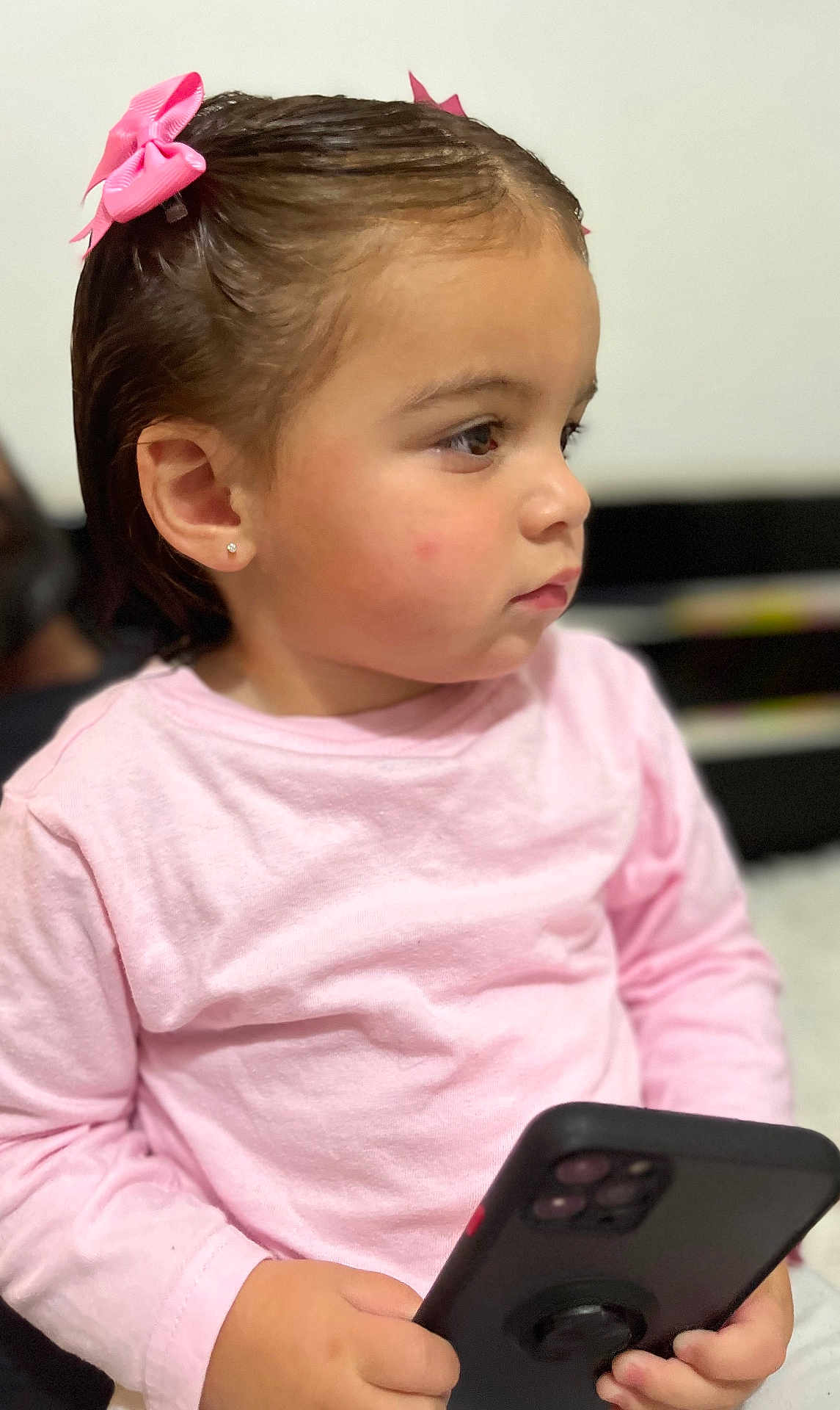 Lissa Kolotian a rejoint le concours — aidez-le/la à gagner de superbes lots ! child, toddler, girl, portrait, profile, pink_shirt, hair_bow, earring, holding_phone, smartphone, sitting, indoor, cute, chubby_cheeks, side_view, focused, blurry_background, casual_clothing, young_child, closeup
