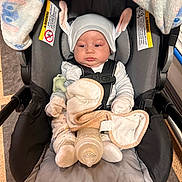 Jeremiah is registered to the contest to win money with this photo: baby, car_seat, blanket, bottle, pacifier, hat, bunny_ears, clothing, infant, child, indoors, seatbelt, soft_toy, flooring, baby_gloves, baby_sock, face, person, portrait, cute