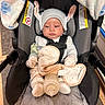 baby, car_seat, blanket, bottle, pacifier, hat, bunny_ears, clothing, infant, child, indoors, seatbelt, soft_toy, flooring, baby_gloves, baby_sock, face, person, portrait, cute