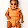 baby, infant, smiling, dimples, curly_hair, bow, orange_onesie, socks, cute, portrait, happy, child, young, studio, white_background, skin, face, person, baby_clothing, sitting