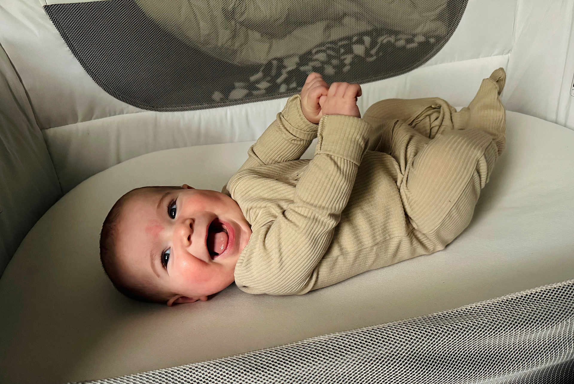 James is registered to the contest to win money with this photo: baby, infant, smiling, joyful, pajamas, cozy, cushion, mesh, crib, lying_down, happy, child, soft_surface, indoors, cute, baby_clothes, hands, feet, face, portrait