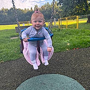 Marnie-Louise is registered to the contest to win money with this photo: baby, child, swing, playground, grass, blanket, pink_bow, jacket, outdoor, daylight, fence, trees, sky, clouds, happy, smiling, person, fun, park, play