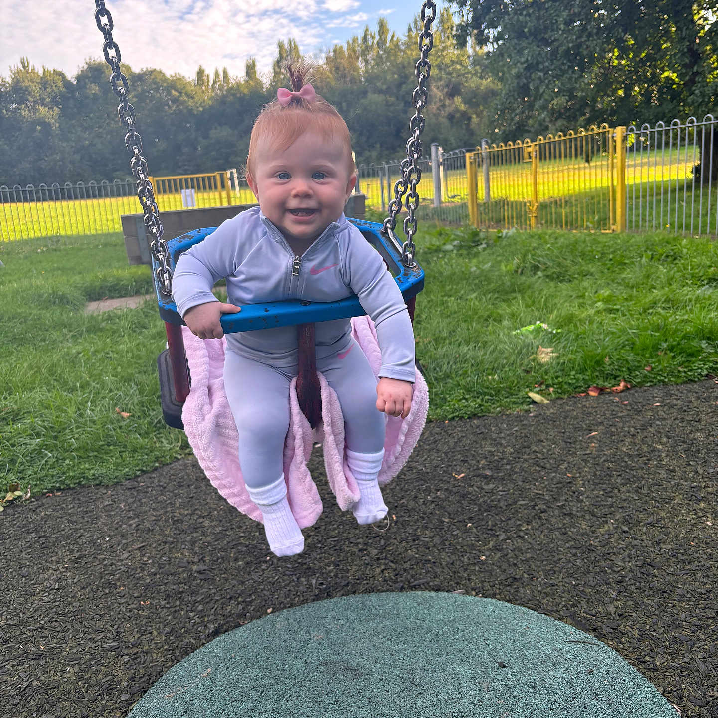 Marnie-Louise is registered to the contest to win money with this photo: baby, blanket, child, clouds, daylight, fence, fun, grass, happy, jacket, outdoor, park, person, pink_bow, play, playground, sky, smiling, swing, trees