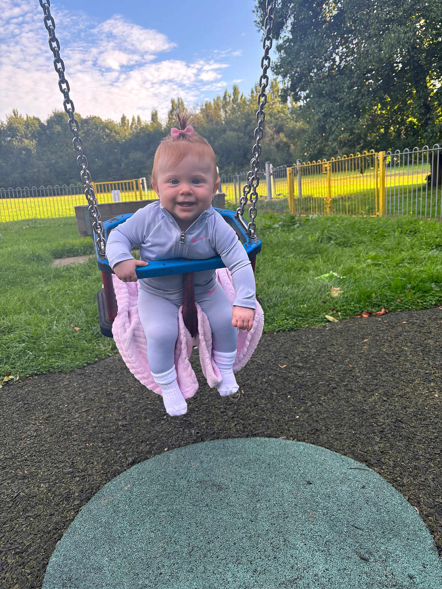 Marnie-Louise is registered to the contest to win money with this photo: baby, child, swing, playground, grass, blanket, pink_bow, jacket, outdoor, daylight, fence, trees, sky, clouds, happy, smiling, person, fun, park, play