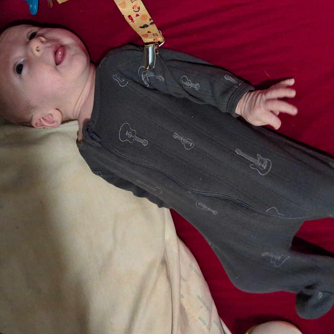 Chance is registered to the contest to win money with this photo: baby, bed, beige_blanket, blanket, child, curious, fabric, footed_pajamas, guitar_print, indoor, infant, lying_down, mouth_open, onesie, person, pillow, red_sheet, sleepwear, small_hand, soft_lighting