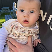 Florence is registered to the contest to win money with this photo: baby, child, infant, person, hand, watch, floral_dress, outdoor, daylight, curious, face, skin, eyes, clothing, adult_hand, picnic_table, blurred_background, garden, sunlight, portrait