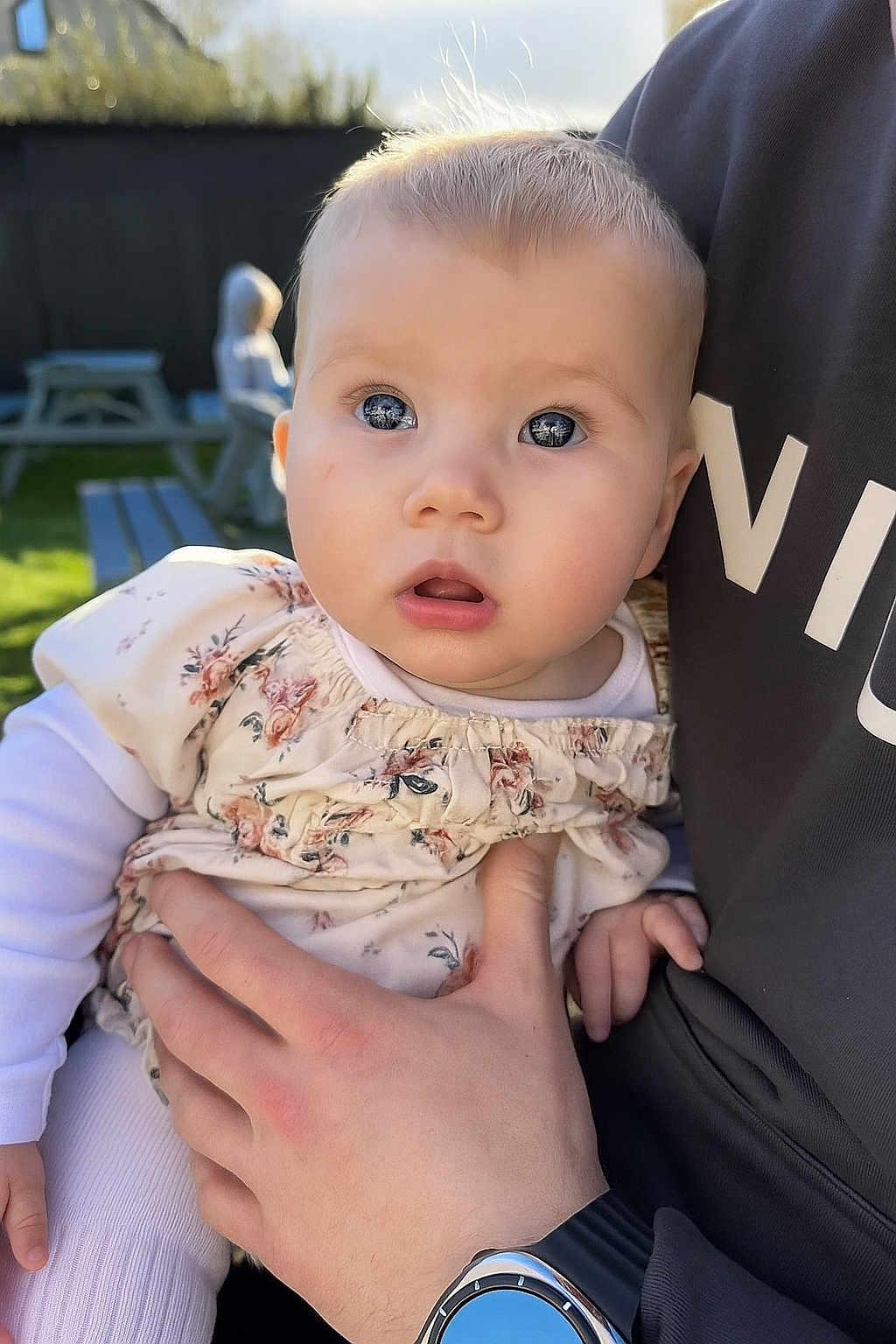 Florence is registered to the contest to win money with this photo: baby, child, infant, person, hand, watch, floral_dress, outdoor, daylight, curious, face, skin, eyes, clothing, adult_hand, picnic_table, blurred_background, garden, sunlight, portrait