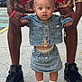 toddler, child, denim_jacket, denim_skirt, sneakers, adult_hands, sidewalk, street, gold_necklace, shorts, floral_pattern, curly_hair, blue_eyes, casual_clothing, urban, holding_hands, outdoor, fashion, expression, person