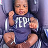 baby, car_seat, child, close_up, clothing, curly_hair, eyes, face, fist, hand, harness, indoor, infant, newborn, portrait, seated, shirt, smiling, socks, vehicle