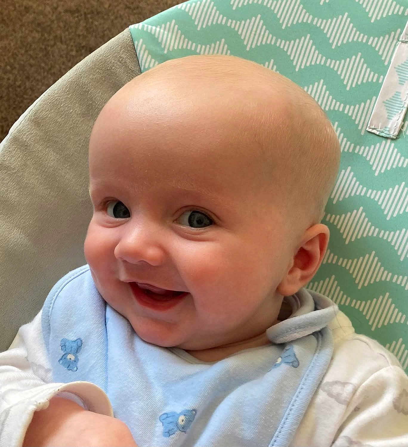 Logan is registered to the contest to win money with this photo: baby, smiling, face, blue_bib, infant, child, cute, patterned_cushion, happy, closeup, skin, eyes, cheeks, clothing, portrait, indoor, baby_bib, baby_clothing, head, expression