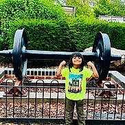 Mifraa is registered to the contest to win money with this photo: child, smiling, flexing, muscles, green_shirt, pink_shoes, outdoor, barbell, weight, fence, leaves, grass, trees, bushes, park, playful, confident, person, casual_clothing, daytime