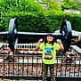 child, smiling, flexing, muscles, green_shirt, pink_shoes, outdoor, barbell, weight, fence, leaves, grass, trees, bushes, park, playful, confident, person, casual_clothing, daytime