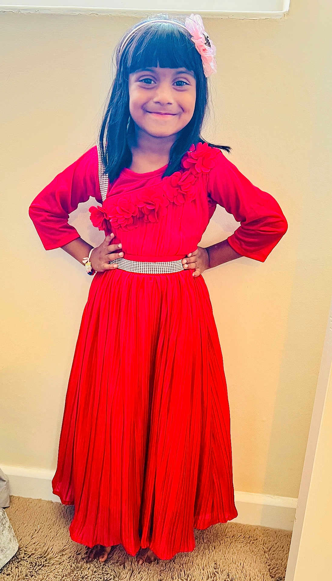 Mifraa is registered to the contest to win money with this photo: child, girl, red_dress, flower_headband, smiling, hands_on_hips, indoor, carpet, wall, long_hair, barefoot, pleated_skirt, flower_decoration, bracelet, portrait, confident, headband, person, standing, happy