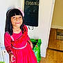 Mifraa is registered to the contest to win money with this photo: child, girl, pink_dress, smiling, indoors, kitchen, apron, flooring, wood_floor, bag, black_apron, headband, flower_accessory, hand, long_hair, posing, wall, doorway, bright_colors, portrait