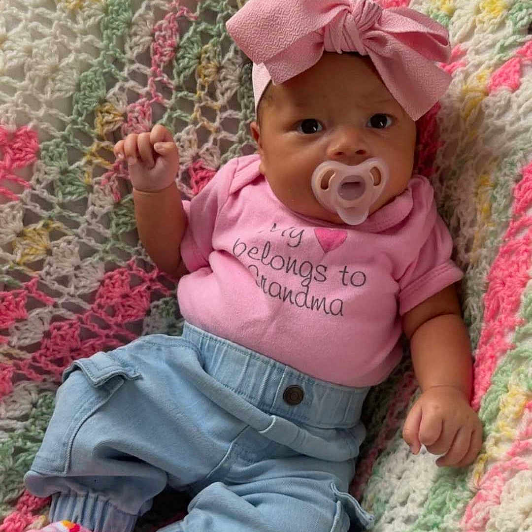 Island is registered to the contest to win money with this photo: baby, blue_pants, child, colorful, cozy, crocheted_blanket, cute, face, hand, headwear, indoor, infant, pacifier, pink_bow, pink_shirt, pink_socks, portrait, seated, soft_texture, text_on_shirt