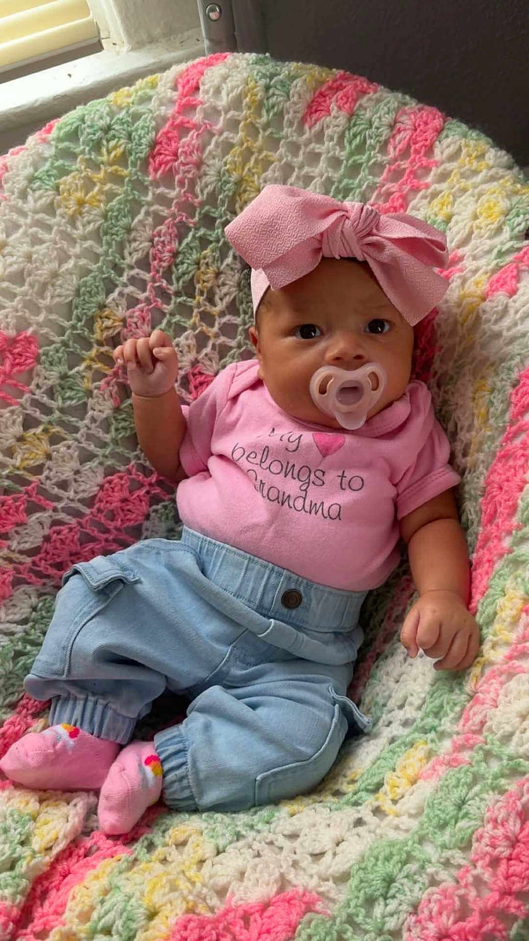 Island is registered to the contest to win money with this photo: baby, pacifier, pink_bow, crocheted_blanket, pink_shirt, blue_pants, pink_socks, infant, child, cute, portrait, indoor, colorful, text_on_shirt, hand, face, headwear, seated, cozy, soft_texture