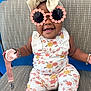 baby, child, sunglasses, flower_pattern, bow_headband, pacifier, bracelet, smiling, seated, chair, patterned_fabric, cute, happy, accessory, fashion, indoors, portrait, cute_outfit, infant, playful