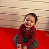 toddler, child, smiling, red_blanket, curly_hair, sitting, barefoot, long_sleeve_shirt, pants, indoor, wooden_wall, happy, cute, portrait, young_child, playful, casual_clothing, bare_feet, person, baby