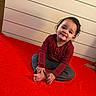 toddler, child, sitting, smiling, red_blanket, long_sleeve_shirt, gray_pants, barefoot, indoors, wood_panel_wall, cute, portrait, young_child, happy, casual_clothing, floor_seating, small_person, playful, natural_light, cozy