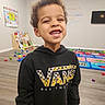 child, smiling, curly_hair, hoodie, vans_logo, playroom, toys, colorful, educational_poster, wooden_floor, happy, indoor, young_kid, casual_clothing, portrait, boy, floor_mat, television, wall_art, bright_lighting