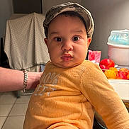 Noah participe au concours pour gagner de l'argent avec cette photo : baby, child, face, person, camo_cap, orange_outfit, kitchen, fruit, bowl, table, arm, bracelet, tile_floor, indoor, curious_expression, plush_outfit, lighting, background, home, support