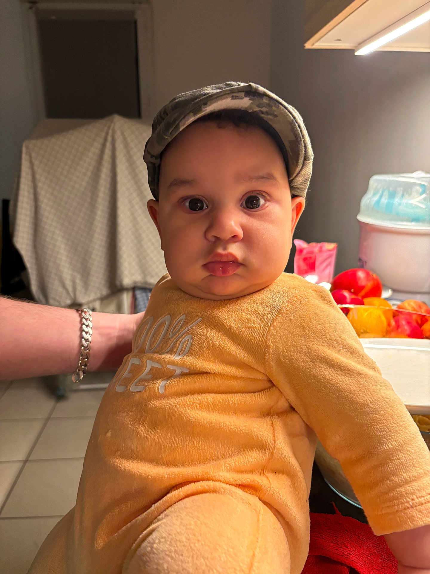 Noah participe au concours pour gagner de l'argent avec cette photo : baby, child, face, person, camo_cap, orange_outfit, kitchen, fruit, bowl, table, arm, bracelet, tile_floor, indoor, curious_expression, plush_outfit, lighting, background, home, support