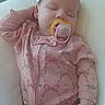 baby, sleeping, pacifier, pink_onesie, bows_pattern, cushion, relaxed, infant, child, peaceful, resting, hand, face, closed_eyes, indoor, soft_lighting, cute, cozy, portrait, person