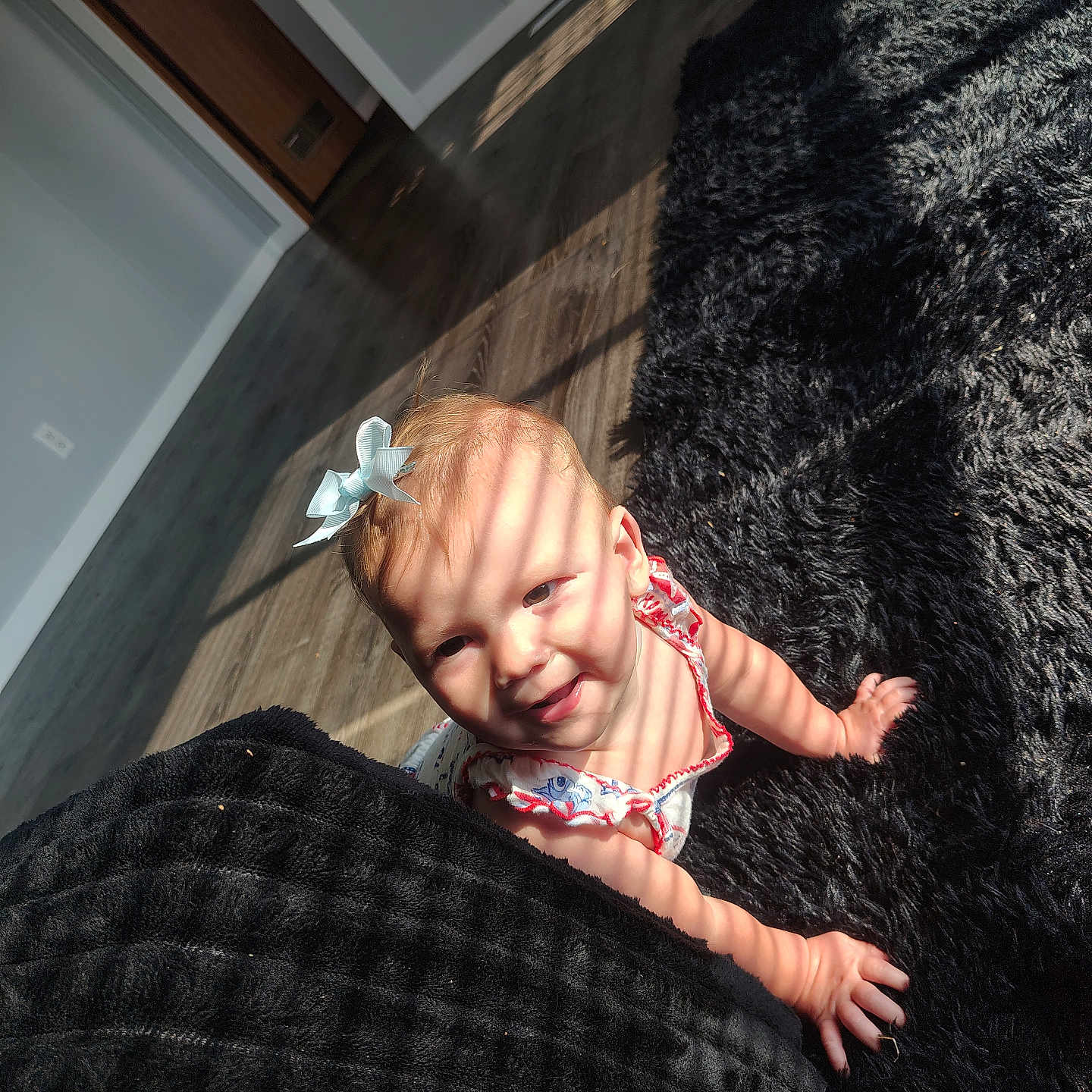 Maia is registered to the contest to win money with this photo: baby, child, crawling, blue_bow, sunlight, shadow, striped_light, indoor, rug, floor, wooden_floor, black_rug, smiling, happy, person, cute, infant, home, playful, face