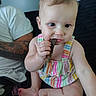 baby, child, infant, person, colorful_clothing, striped_outfit, sitting, indoor, tattoo, adult, hand, face, foot, curious, munching, black_object, candid, portrait, skin, expression