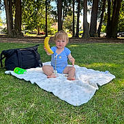 Asher is registered to the contest to win money with this photo: banana, black_bag, blanket, blue_onesie, child, daytime, grass, green_bottle, happy, nature, outdoor, pacifier, park, playful, sitting, summer, sunlight, toddler, trees, young_child