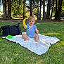 banana, black_bag, blanket, blue_onesie, child, daytime, grass, green_bottle, happy, nature, outdoor, pacifier, park, playful, sitting, summer, sunlight, toddler, trees, young_child