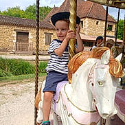 Philemon a rejoint le concours — aidez-le/la à gagner de superbes lots ! child, boy, carousel, horse, ride, hat, striped_shirt, shorts, outdoor, daytime, grass, stone_building, wooden_door, greenery, playground, amusement_ride, smile, fun, summer, vacation