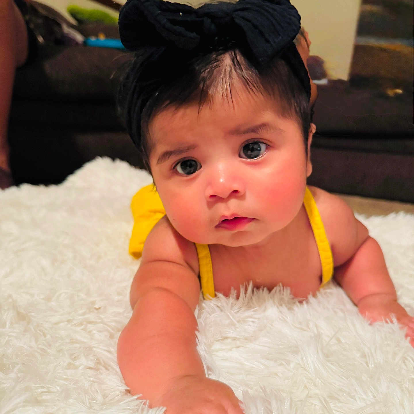 Tessa Kaeli joined the competition — help win amazing prizes! baby, child, infant, headband, black_bow, yellow_clothing, fluffy_rug, white_rug, indoor, living_room, carpet, couch, person, cute, curious, reaching, skin, face, portrait, toddler