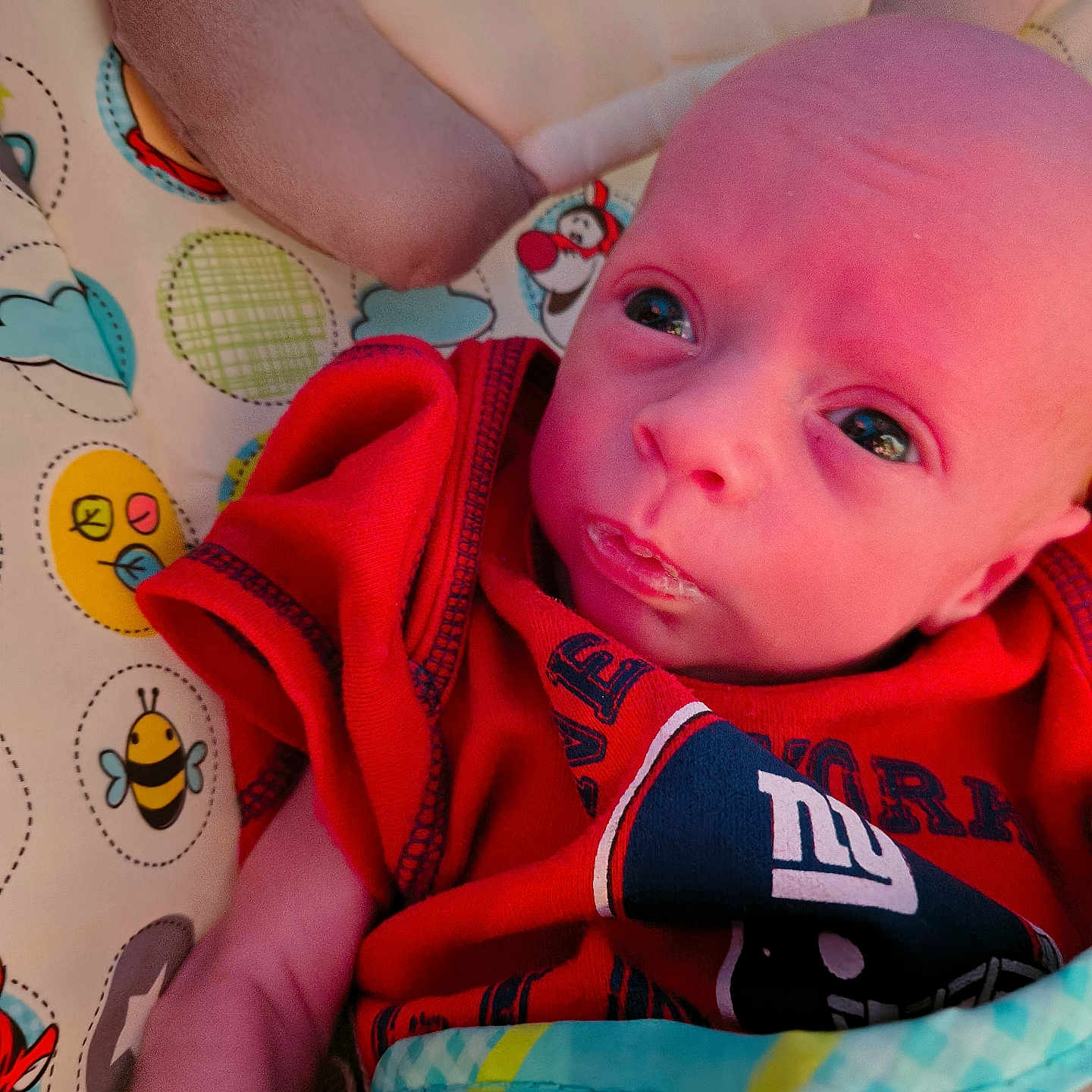 Dylan is registered to the contest to win money with this photo: baby, infant, close_up, face, red_clothing, blanket, patterned_blanket, cartoon, toy, soft_toy, blue, yellow, pink_skin, eyes, expression, newborn, cute, child, portrait, lying_down