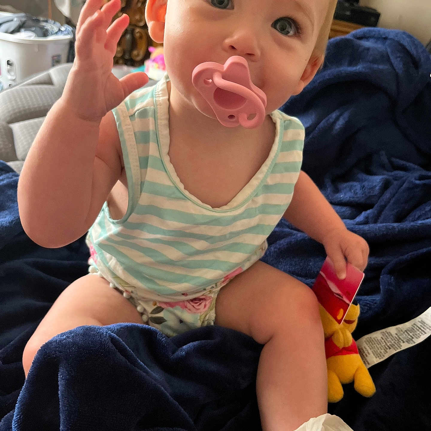 Teagan is registered to the contest to win money with this photo: baby, blanket, blue_blanket, child, curious, cute, diaper, face, hand_raised, home, indoor, infant, pacifier, person, plush_toy, sitting, smile, soft_toy, striped_clothing, toy