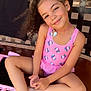 child, girl, smiling, curly_hair, pink_swimsuit, unicorn_pattern, sitting, bench, outdoor, playful, necklace, barefoot, toy_car, tile_floor, casual, backyard, relaxed, portrait, happy, summer