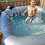 activity, backyard, boy, child, daytime, fun, greenery, inflatable_pool, outdoor, person, playful, pool, recreation, splash, summer, sunlight, swimwear, water, water_droplets, wooden_fence