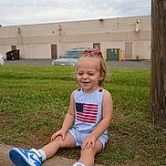 Blakely joined the competition — help win amazing prizes! toddler, child, sitting, outdoor, grass, curb, sneakers, blue_shoes, american_flag, clothing, happy, smiling, shorts, hair_bow, red_bow, concrete, building, car, pole, daytime