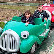 Kane is registered to the contest to win money with this photo: child, children, amusement_park, ride, toy_car, green_car, red_car, outdoor, track, trees, grass, jacket, happy, play, fun, person, pavement, daytime, smile, young