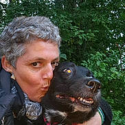 Belle participe au concours pour gagner de l'argent avec cette photo : person, dog, black_dog, collar, outdoor, greenery, trees, close_up, affection, kiss, short_hair, gray_hair, pet, animal, nature, face, hand, jacket, scarf, flash_reflection