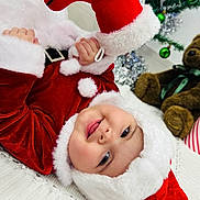 Maria joined the competition — help win amazing prizes! baby, child, santa_claus_costume, red_clothing, white_fur, smiling, face, teddy_bear, toy, holiday, christmas_tree, ornaments, blanket, indoor, floor, festive, cute, person, headwear, happy
