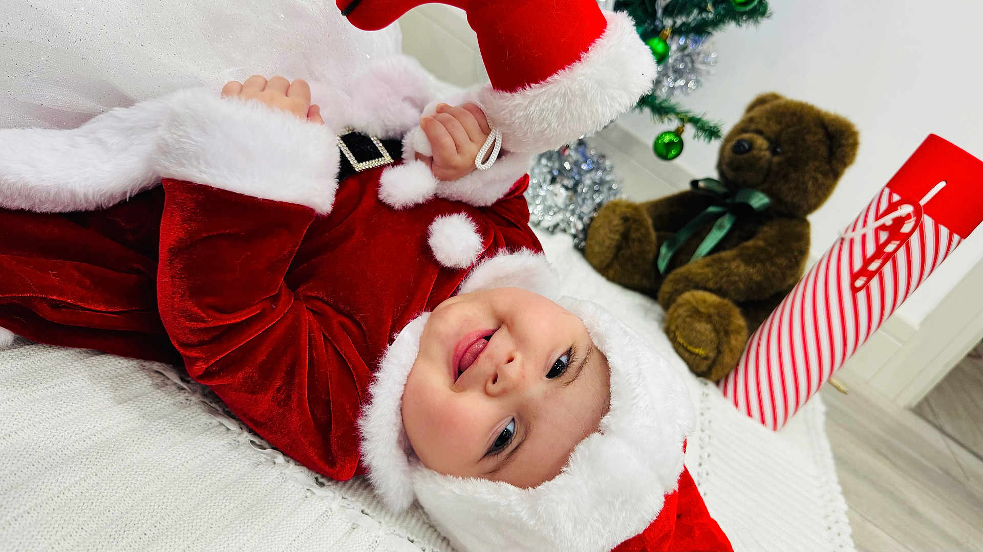 Maria joined the competition — help win amazing prizes! baby, child, santa_claus_costume, red_clothing, white_fur, smiling, face, teddy_bear, toy, holiday, christmas_tree, ornaments, blanket, indoor, floor, festive, cute, person, headwear, happy