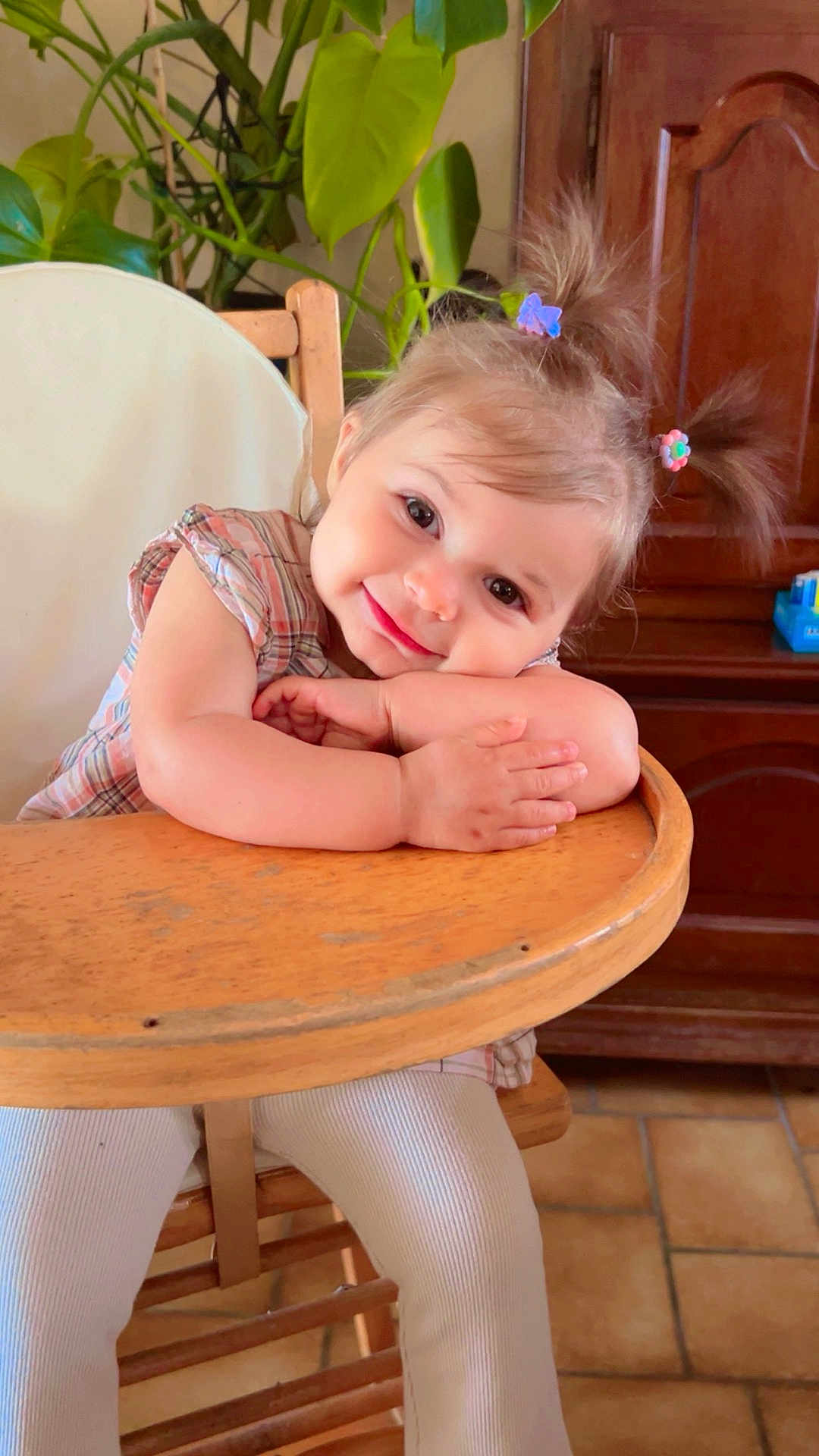 Mayhanna participe au concours pour gagner de l'argent avec cette photo : child, cozy, cute, face, floor_tiles, hair_clips, hands, happy, highchair, home, indoor, person, pigtails, plaid_shirt, plant, portrait, smiling, toddler, white_pants, wooden_furniture