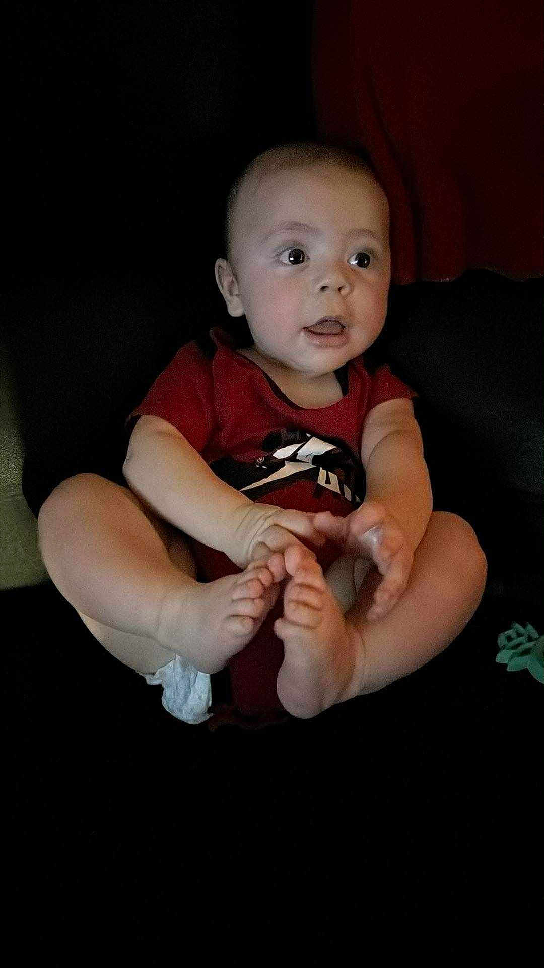Xavier joined the competition — help win amazing prizes! baby, child, infant, red_onesie, feet, hands, face, curious, sitting, indoor, person, skin, expression, toes, young, cute, portrait, dark_background, candid, eyes