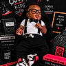 baby, child, glasses, bow_tie, suspenders, sneakers, black_clothing, chalkboard, books, mug, backpack, pencil, lamp, composition_book, text, indoor, portrait, cute, smiling, props