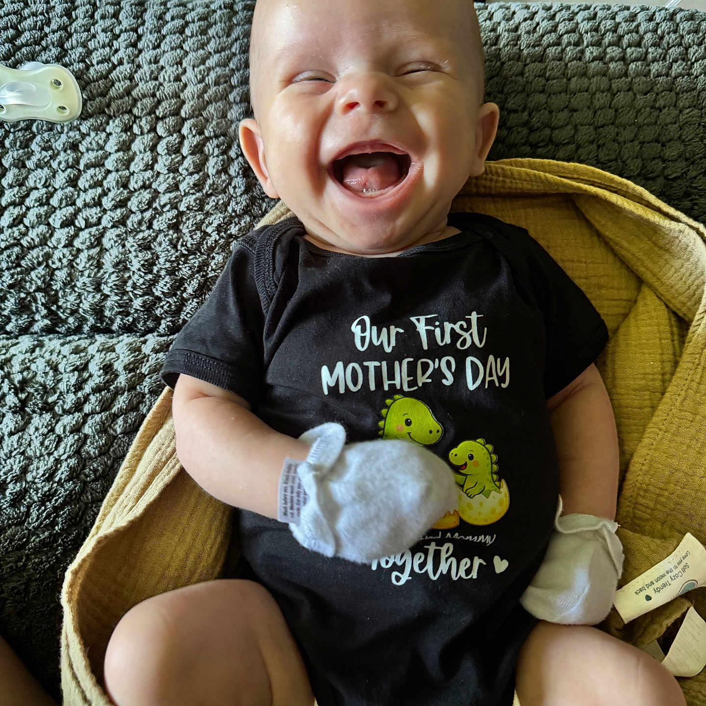 Waylon joined the competition — help win amazing prizes! baby, infant, smiling, laughing, black_onesie, blanket, yellow_blanket, mittens, cute, happy, face, person, indoors, celebration, mother's_day, dinosaur_graphic, clothing, texture, cozy, portrait
