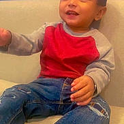 Aarav joined the competition — help win amazing prizes! toddler, child, sitting, couch, red_sweatshirt, gray_sleeves, ripped_jeans, blue_background, happy, curious, casual_clothing, indoor, young_child, smiling, cozy, comfortable, feet_up, portrait, person, casual
