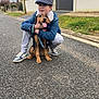 Hugo a rejoint le concours — aidez-le/la à gagner de superbes lots ! boy, child, dog, pet, street, sidewalk, tree, house, cap, denim_jacket, gray_pants, sneakers, leash, grass, suburban, outdoor, sky, cloudy, happy, crouching