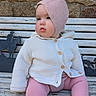 baby, child, pink_bonnet, white_cardigan, knitwear, bench, wooden_bench, stone_wall, outdoor, pensive, cute, portrait, face, sitting, cozy, winter_clothing, plush, infant, candid, soft_lighting