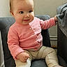 baby, child, infant, pink_cardigan, beige_pants, socks, chair, gray_chair, blanket, indoor, wall, curly_hair, cute, smiling, seated, furniture, cozy, small_hands, looking_away, soft_lighting